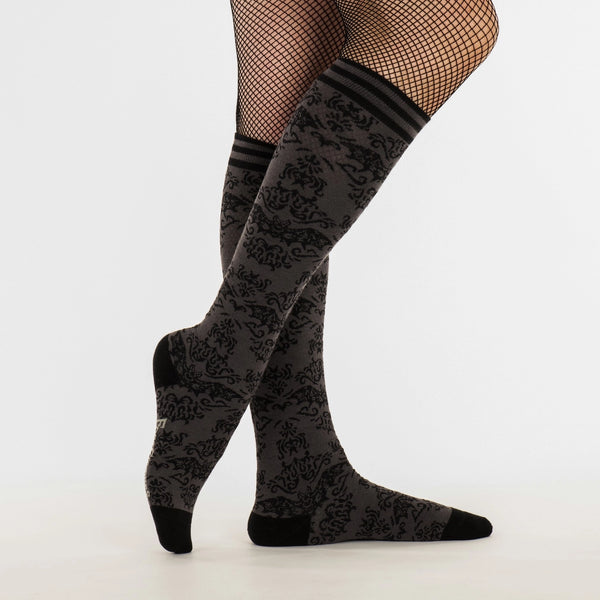 Knee socks with a monochromatic damask pattern of flying bats. Striped grey and black bands at the top of each sock and solid black toes and heels. Shown on a model wearing fishnet tights underneath the socks, shown from the side