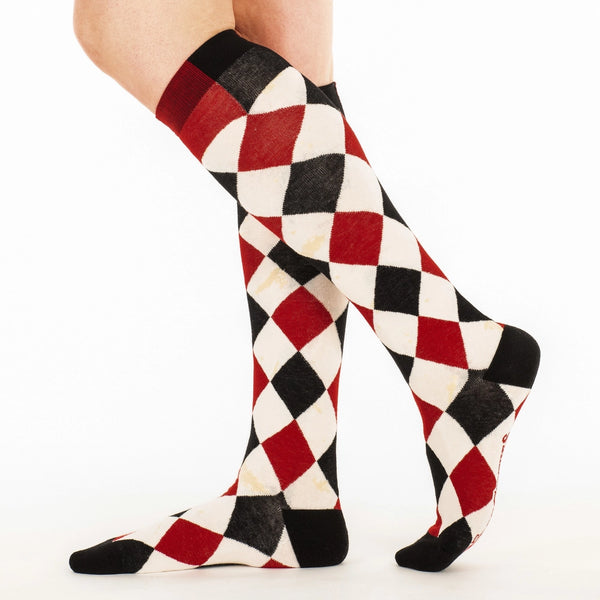 knee socks in a black, red, and cream colored harlequin pattern- with faux-antiqued yellowed age spots throughout each cream colored diamond in the pattern. Shown on model from the side 