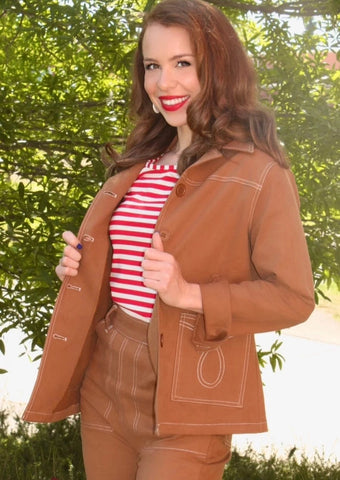 Model wearing a 1950s style button down jean jacket in a toasty cashew brown color. It has matching brown plastic buttons down the chest and at the cuffs and white top stitching detail in a loop shape on each patch pocket