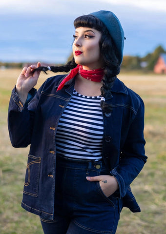 Model wearing a 1950s style button down jean jacket in an indigo blue color. It has matching dark navy blue plastic buttons down the chest and at the cuffs and denim yellow top stitching detail in a loop shape on each patch pocket