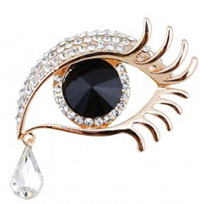 Gold metal brooch of an eye with eyelashes and a large black faceted jewel for an iris. There is a teardrop hanging from the corner of the eye made of a tear shaped faceted jewel