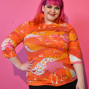 A plus size model wearing a cotton 3/4 sleeve knit top with an all-over pattern that includes pinwheels, apples, hands, trees, frogs, and cows. It is printed in shades of red, orange, pink, and white. Shown from the front