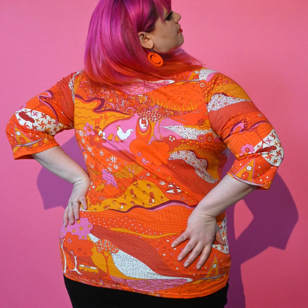 A plus size model wearing a cotton 3/4 sleeve knit top with an all-over pattern that includes pinwheels, apples, hands, trees, frogs, and cows. It is printed in shades of red, orange, pink, and white. Shown from the back 
