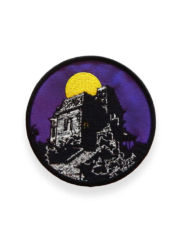 Round embroidered patch picturing a haunted house seen in silhouette in front of a dark purple sky and full yellow moon. With a black border