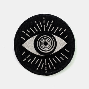 Round embroidered patch of an abstract illustration of an eye with a swirly round iris in silvery white. Surrounded by matching lines and on a black background 