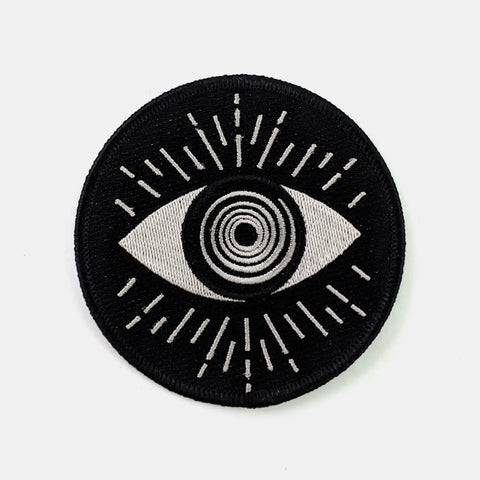 Round embroidered patch of an abstract illustration of an eye with a swirly round iris in silvery white. Surrounded by matching lines and on a black background 
