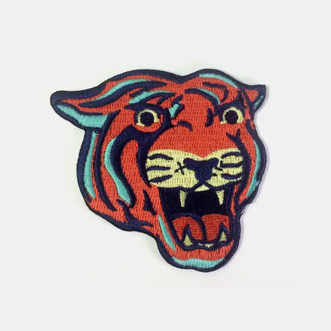 Embroidered patch of a tiger’s head with its mouth open snarling. In shades of coral, bright teal, navy blue, and yellow
