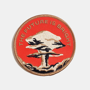 Round embroidered patch of a brown, beige, and black mushroom cloud with the message “THE FUTURE IS BRIGHT” in brown on a red background. Brown border on patch