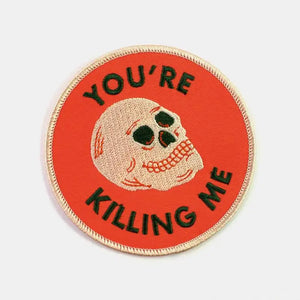 A round embroidered patch with an orange background and cream colored border. Shows an illustration of a cream colored skull with forest green and orange details. With the message “YOU’RE KILLING ME” written in forest green capital letters.