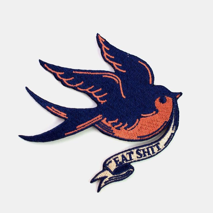 Embroidered patch of a sparrow in traditional tattoo style in coral and navy blue holding a white banner that says “EAT SHIT”