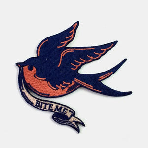 Embroidered patch of a traditional tattoo style sparrow in navy blue and coral holding a banner in its beak reading “BITE ME” in capital letters 