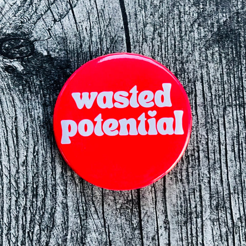 Round metal button with bright red background and “wasted potential” written in white lower case font with a heart dotting the letter i 