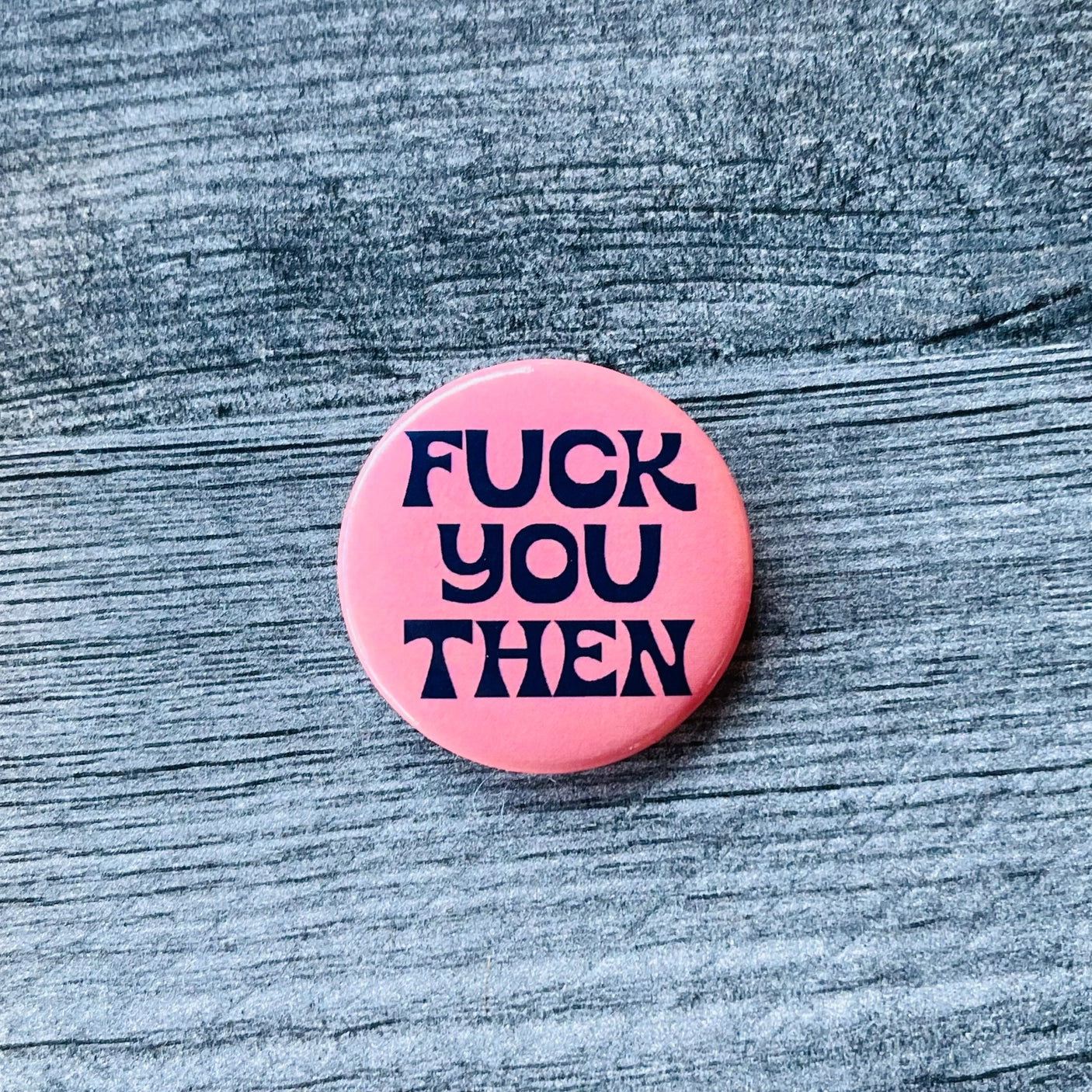 A 1 1/2” round button with a light pink background and the phrase “FUCK YOU THEN” in a curvy retro navy blue font