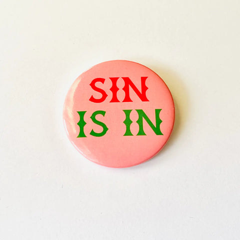 2 1/4” round button with a light pink background and the phrase “SIN IS IN” written in a geometric font in red and green