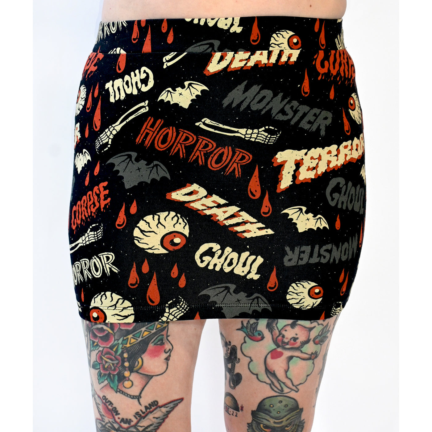A black stretch knit mini skirt with an all-over pattern of bats, skeleton hands, dripping red blood, and eyeballs with the words “DEATH”, “GHOUL”, “MONSTER”, “HORROR” written in vintage movie poster style fonts in red, grey, and beige yellow. Seen up close