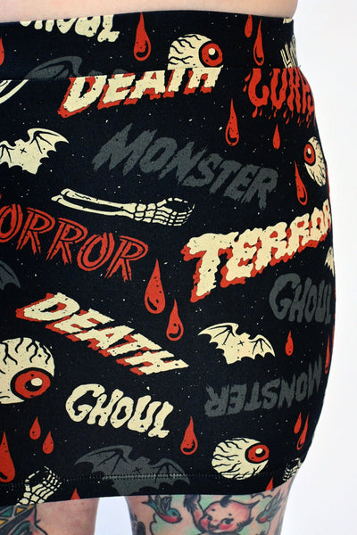 A black stretch knit mini skirt with an all-over pattern of bats, skeleton hands, dripping red blood, and eyeballs with the words “DEATH”, “GHOUL”, “MONSTER”, “HORROR” written in vintage movie poster style fonts in red, grey, and beige yellow. Seen on a model from the waist down in close up