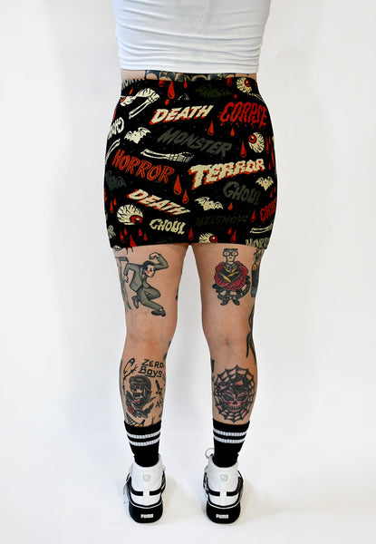 A black stretch knit mini skirt with an all-over pattern of bats, skeleton hands, dripping red blood, and eyeballs with the words “DEATH”, “GHOUL”, “MONSTER”, “HORROR” written in vintage movie poster style fonts in red, grey, and beige yellow. Seen on a model from the waist down from the back