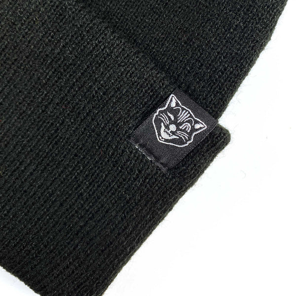 black knit hat with a set of pointy little matching cat ears on top and a rolled cuff. Shown up close with Sourpuss logo brand tag