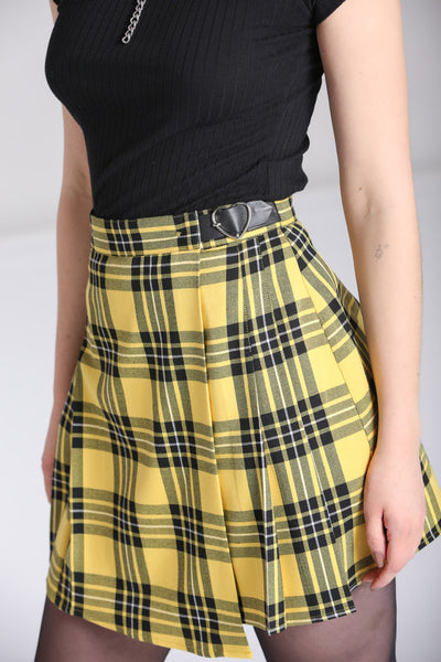 Model wearing a black and yellow tartan print pleated miniskirt with a faux wrap detail in the front. It has a black faux leather strap at the left hip of the waistband with a shiny silver heart shaped buckle. Shown up close from a three quarter angle to show buckle detail 