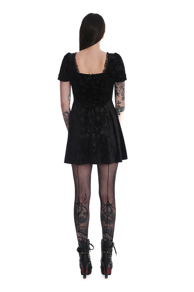 Model wearing a black stretch velvet mini dress with an all over embossed damask pattern of chandeliers, roses, candles, and crescent moons. It has lace trim at the bust and shoulders of the square neckline and a lace up notched detail at the bust. The skirt hits above the knee. Shown from the back