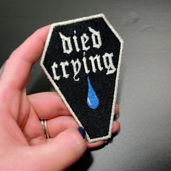 “ died crying” in a gothic font with a big old blue teardrop on an embroidered black coffin shaped patch with white border. Shown held
