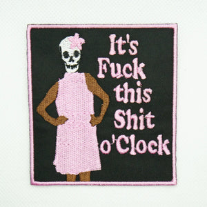 skull headed being in a pink dress with hands on their hips on a black cotton twill background with a matching pink embroidered border with the words “It’s Fuck This Shit o’Clock” in matching pink