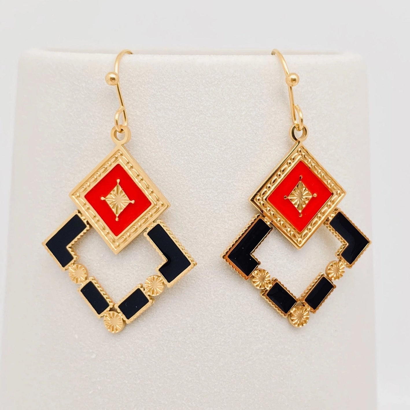 shiny gold metal dangle earrings in the shape of two overlapping squares with small sunburst detailing and black & rich red enameled panels