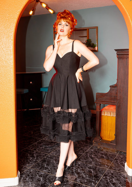 Model wearing black Bengaline sleeveless dress with adjustable straps, exaggerated sweetheart neckline, princess seaming, and a full swing skirt ending in matching black tulle with two strips of ruffled tulle running horizontally along the bottom. Seen from the front 