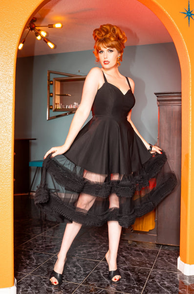 Model wearing black Bengaline sleeveless dress with adjustable straps, exaggerated sweetheart neckline, princess seaming, and a full swing skirt ending in matching black tulle with two strips of ruffled tulle running horizontally along the bottom. Seen from the front with tulle layer held taut