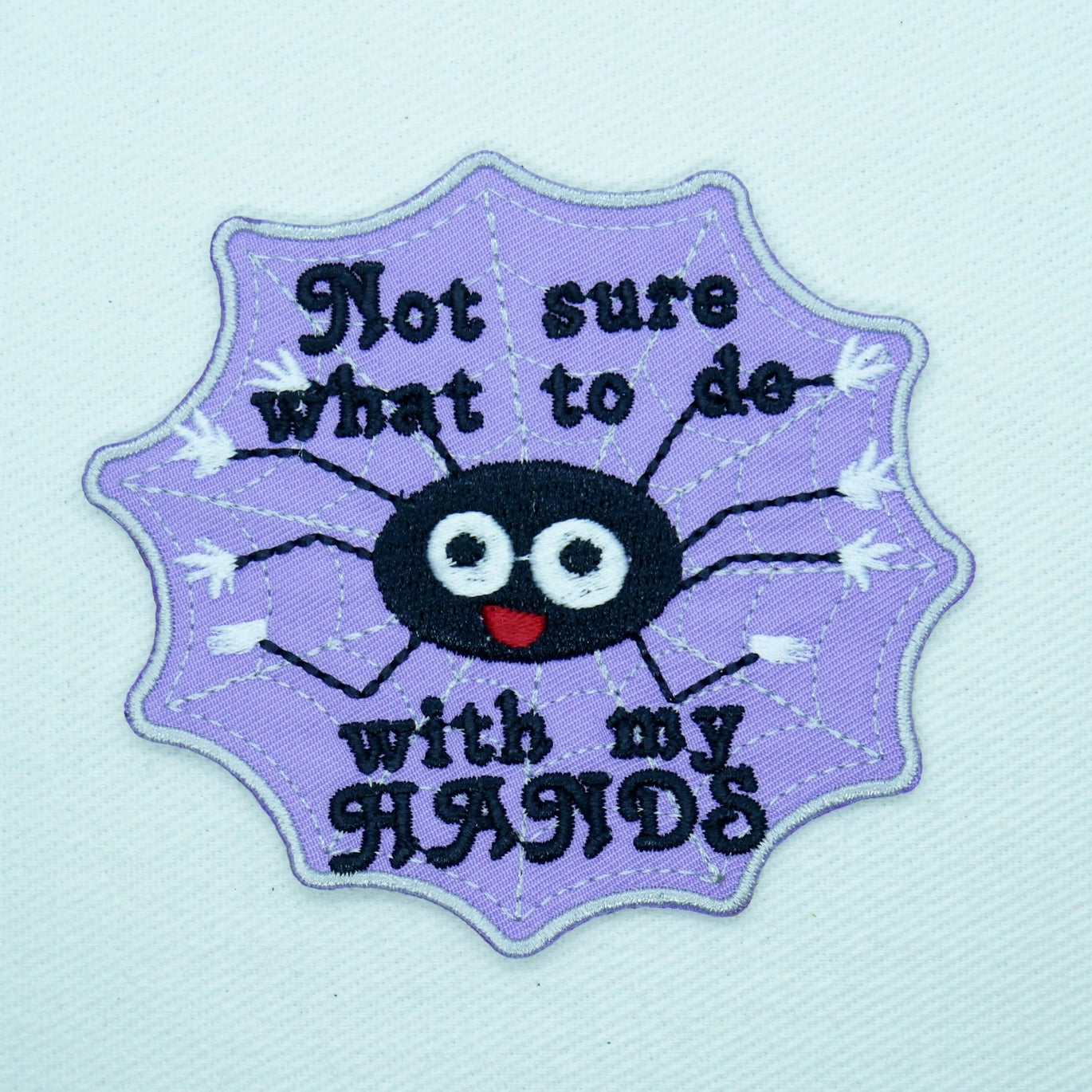 Embroidered purple twill patch of black smiling spider with 8 legs on a web and caption “Not sure what to do with my HANDS” in black 