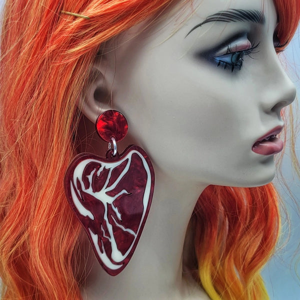 A pair of laser cut acrylic drop earrings in the shape of a rare ribeye steak with marbled red acrylic and two sunny side up eggs. Steak is shown on the ear of a mannequin 