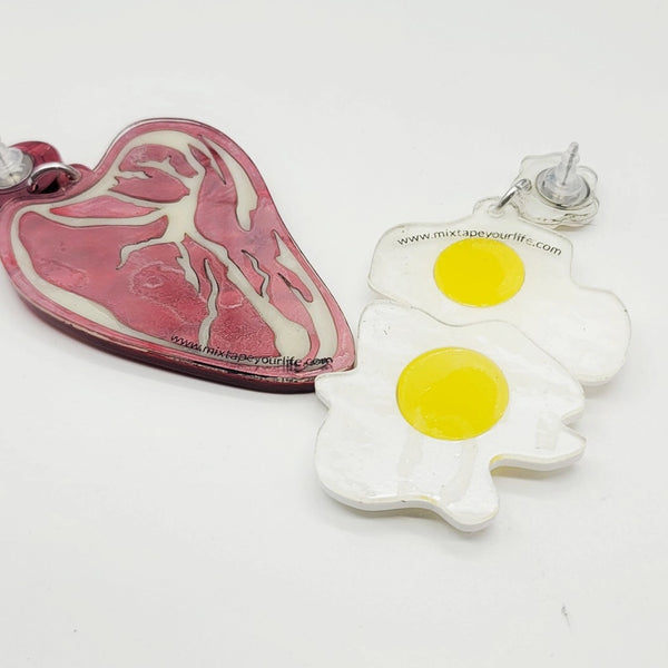 A pair of laser cut acrylic drop earrings in the shape of a rare ribeye steak with marbled red acrylic and two sunny side up eggs. Shown from the back on a white background