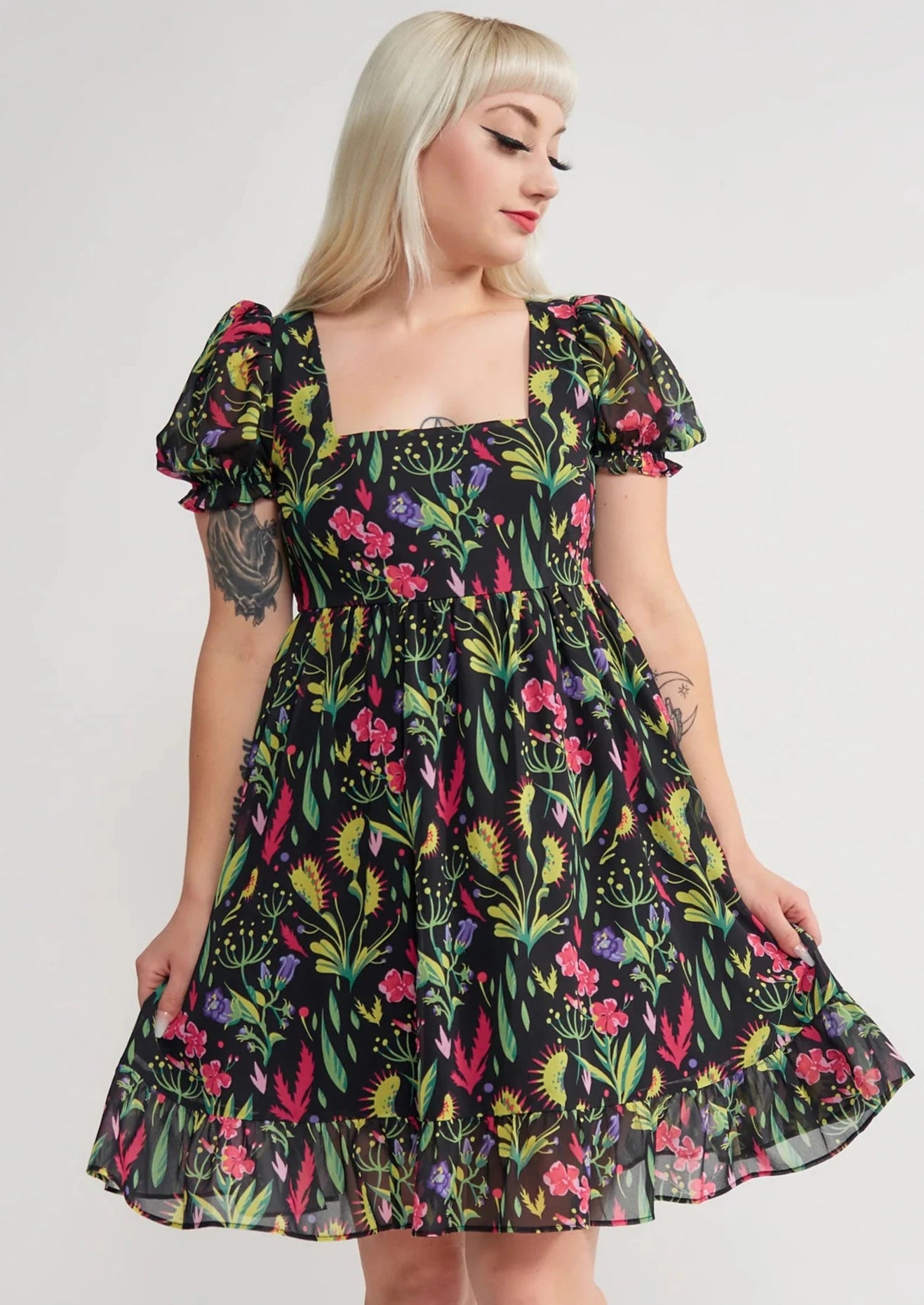 Model wearing a mini dress with a botanical pattern including Venus Flytraps on a black background. It has a square neckline, slightly puffed short sleeves, and a full skirt with a ruffled hem. Shown from the front in close up