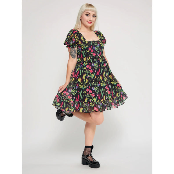 Model wearing a mini dress with a botanical pattern including Venus Flytraps on a black background. It has a square neckline, slightly puffed short sleeves, and a full skirt with a ruffled hem. Shown from a three quarter angle
