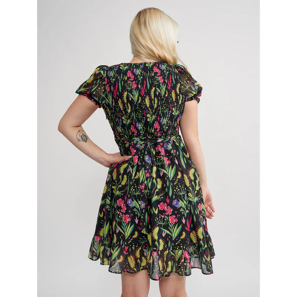 Model wearing a mini dress with a botanical pattern including Venus Flytraps on a black background. It has a square neckline, slightly puffed short sleeves, and a full skirt with a ruffled hem. Shown from behind 