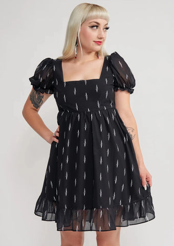 Model wearing a black mini dress with an all-over tiny pattern of white traditional tattoo style daggers. It has a square neckline, slightly puffed short sleeves, and a full skirt with a ruffled hem. Shown from the front in close up