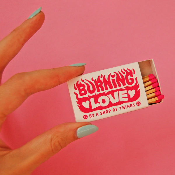 Cardboard box of 18 safety matches in a screen printed box with “BURNING LOVE” “BY A SHOP OF THINGS” written among illustrated flames in bright pink. Shown from front