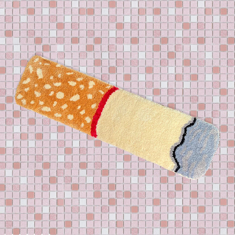 Rectangular rug in the shape of a cigarette with ash and spotted brown filter. Shown flat on a pink tile floor