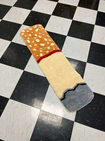 Rectangular rug in the shape of a cigarette with ash and spotted brown filter. Shown on a black and white checkered linoleum floor