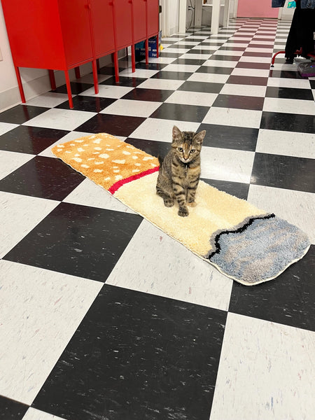 Rectangular rug in the shape of a cigarette with ash and spotted brown filter. Shown on a black and white checkered linoleum floor. Cat standing on rug for scale