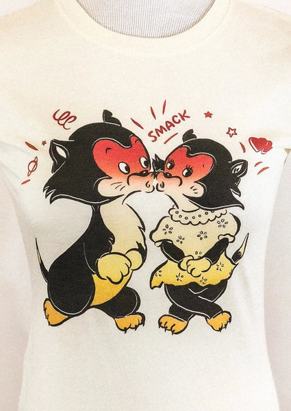 Warm off-white fitted cotton t-shirt with a vintage valentine style illustration of a pair of black and white cats with red blushing faces surrounded by hearts, lines, and “SMACK” as onomatopoeia. Shown on a dress form in close up