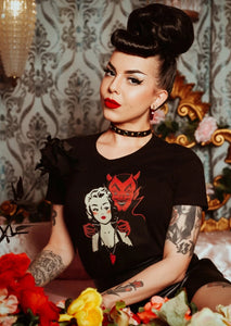 A model wearing a fitted black cotton t shirt with a retro illustration of a devil putting a heart shaped necklace around the neck of a pinup style woman with blonde hair