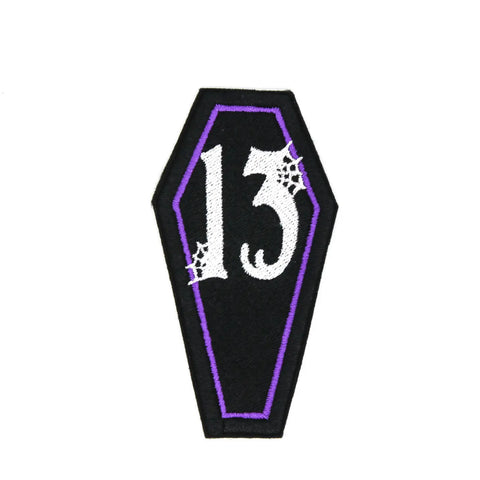 Black embroidered felt patch of a coffin with a purple border and the number 13 written in white in a Gothic font surrounded by spiderwebs 