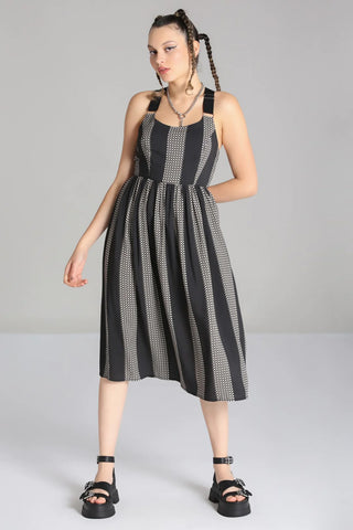 A model wearing a sleeveless dress in a grey and black checker plaid pattern with alternating wide black vertical straps. It has wide adjustable self straps with O-ring detail connecting them to the dress. The dress has princess seaming, a shallow scoop neckline, and a full gathered skirt that ends below the knee. Shown from the front
