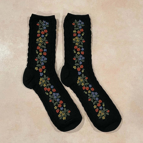 Black cotton socks with both a textured cable knit-in pattern and knit-in pattern of tiny strawberries and yellow & blue flowers. Shown lying flat