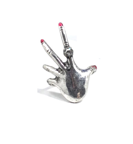 Silver metal with slight burnished finish ring in the shape of a left hand with long fingers, red shiny polish on the nails, and a ring with a clear rhinestone on the index finger. Shown from the front