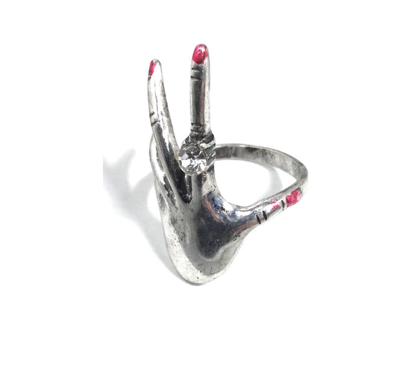 Silver metal with slight burnished finish ring in the shape of a left hand with long fingers, red shiny polish on the nails, and a ring with a clear rhinestone on the index finger. Shown from the side to show band