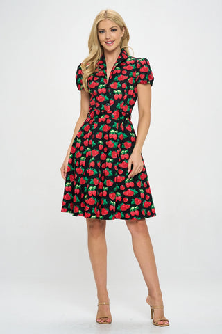 Model wearing a cotton short sleeve flared dress with a notched v-neck collar, princess seaming, puffed cap sleeves, a self removable sash belt, and a flared above the knee skirt. Dress is black with a red and green strawberry pattern. Shown from the front