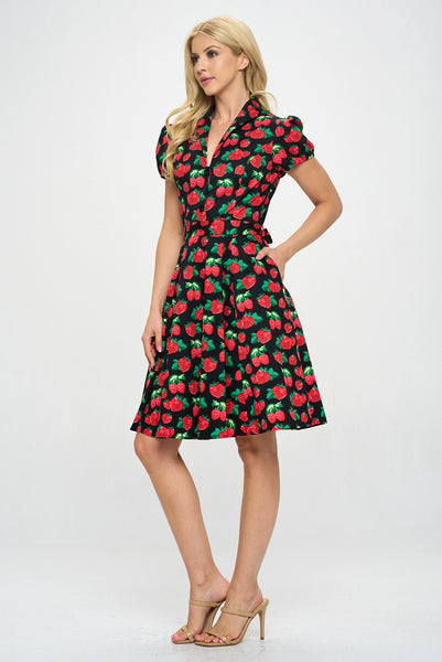 Model wearing a cotton short sleeve flared dress with a notched v-neck collar, princess seaming, puffed cap sleeves, a self removable sash belt, and a flared above the knee skirt. Dress is black with a red and green strawberry pattern. Shown from the side at a three quarter angle 