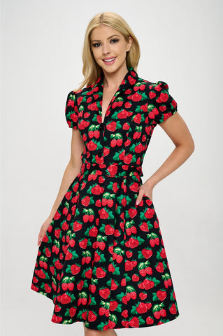 Model wearing a cotton short sleeve flared dress with a notched v-neck collar, princess seaming, puffed cap sleeves, a self removable sash belt, and a flared above the knee skirt. Dress is black with a red and green strawberry pattern. Shown from the front in close up